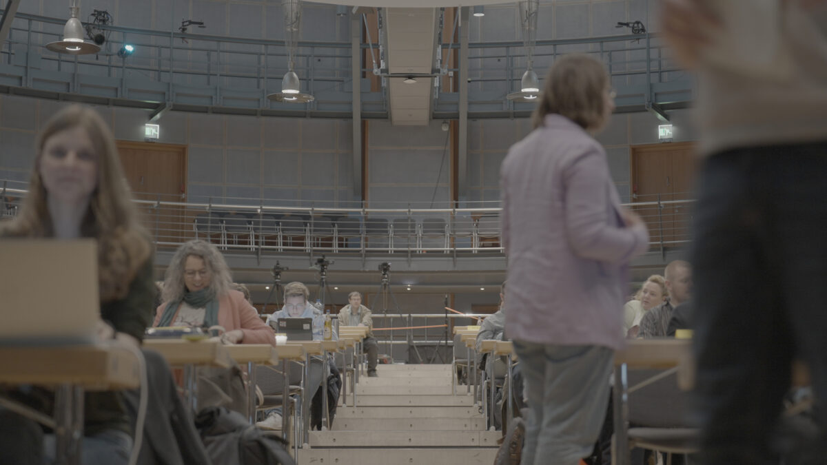 Landespartei 2023 in Celle. Mittelgang Veranstaltungssaal. Personen sitzen an tischen. Vereinzelt bewegen sich Menschen durch den Raum.